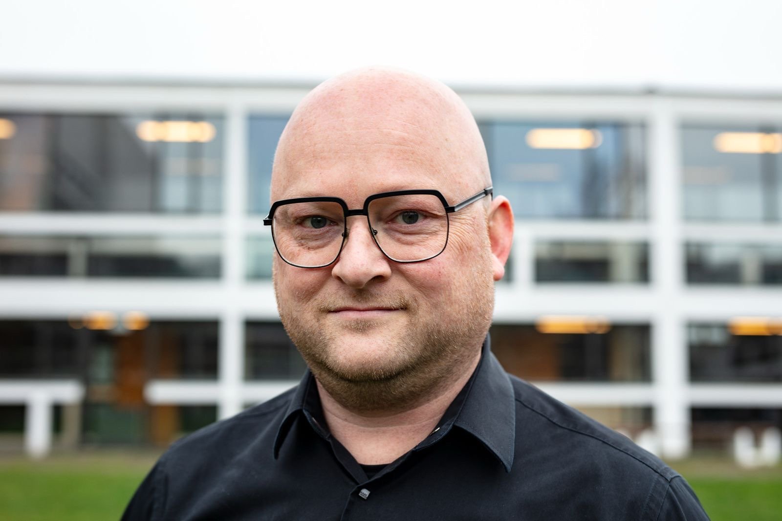 A bald man with glasses wearing a black shirt standing outdoors with a modern building in the background.