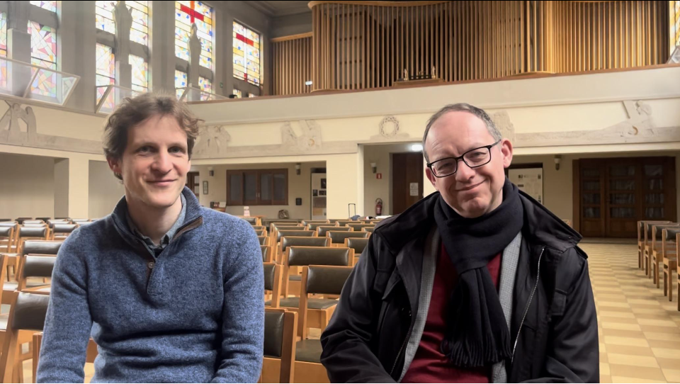 Two men sitting inside a church or chapel, with pews and stained glass windows in the background.
