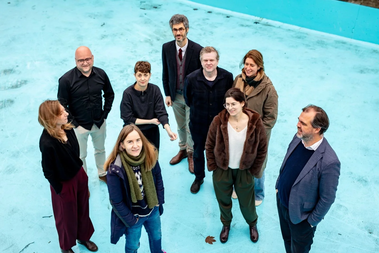 Group of ten diverse people standing and sitting in an outdoor area with a turquoise surface, some looking up at the camera.