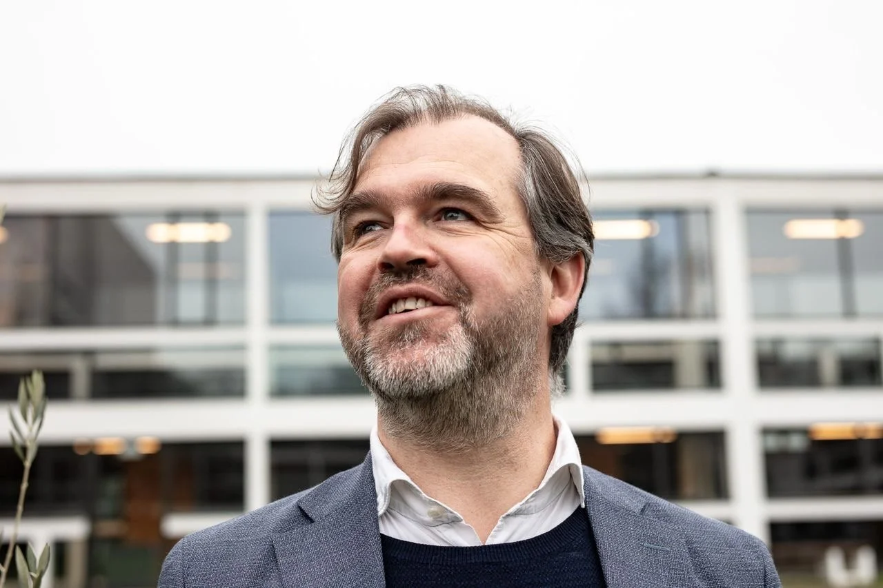 A middle-aged man with gray hair and beard smiling outdoors in front of a modern office building.