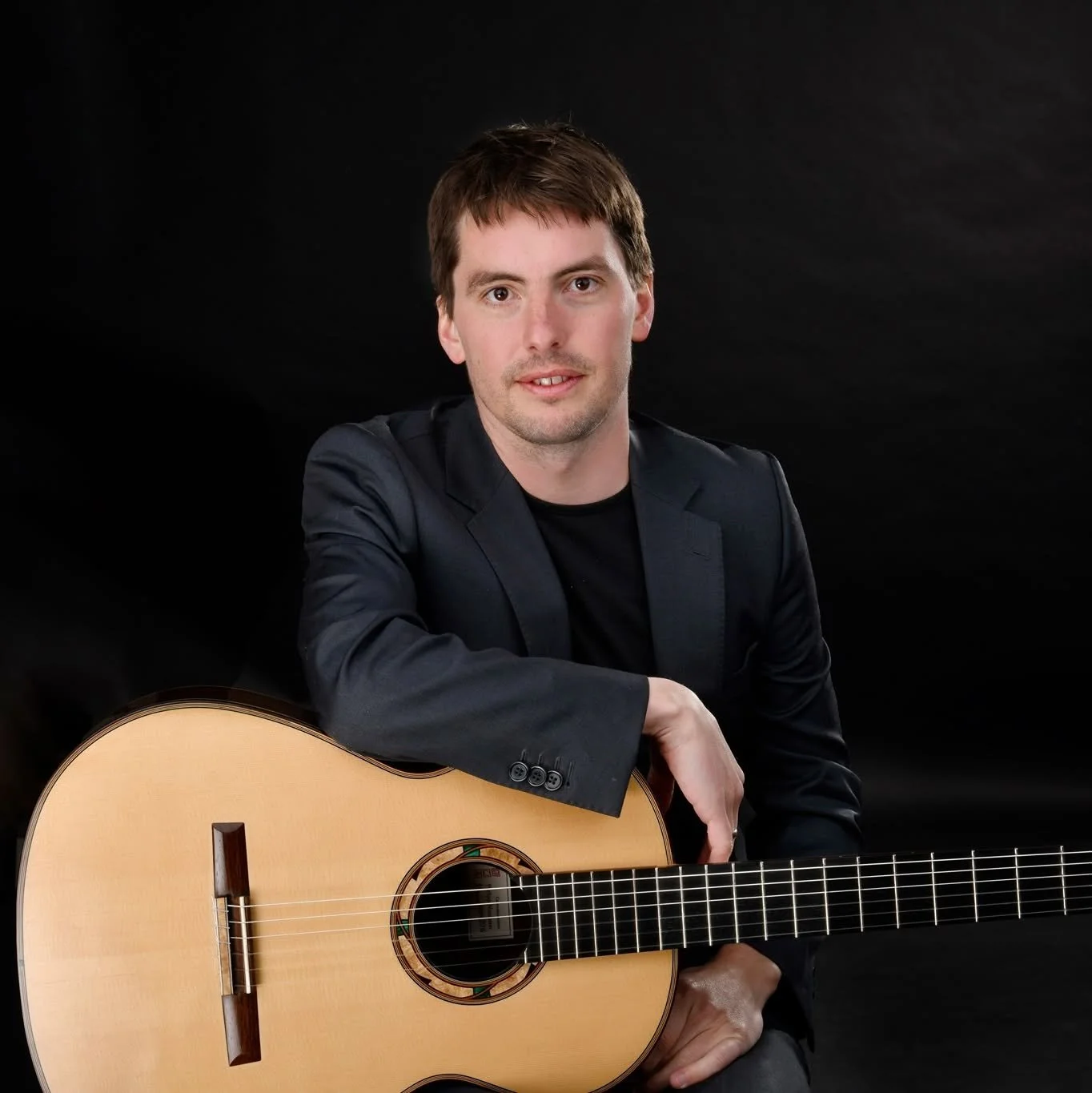 A man with brown hair wearing a black blazer and black shirt, sitting while holding an acoustic guitar, against a black background.