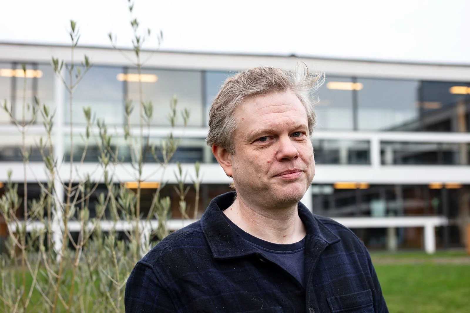A middle-aged man with blonde hair and wearing a dark plaid shirt stands outdoors in front of a modern glass building and some young trees.