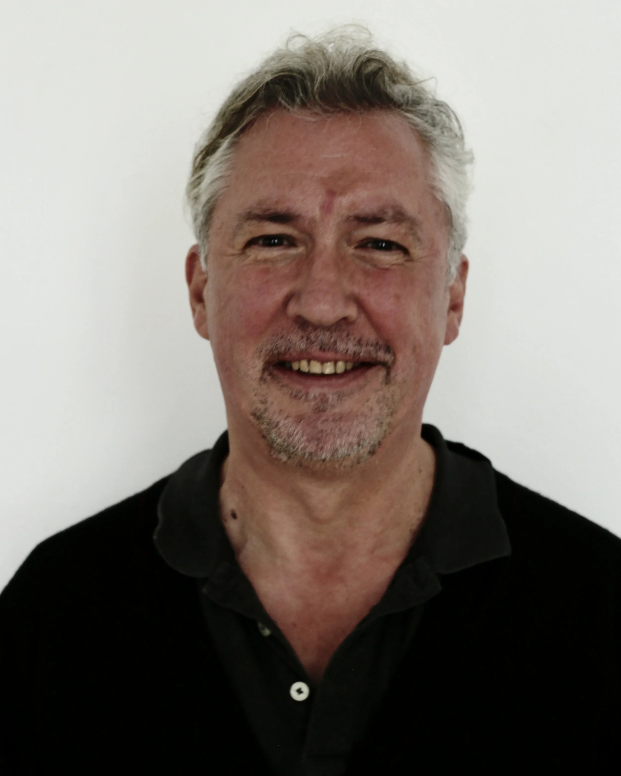 A middle-aged man with gray hair and a goatee smiling at the camera, wearing a black collared shirt, standing against a plain white wall.