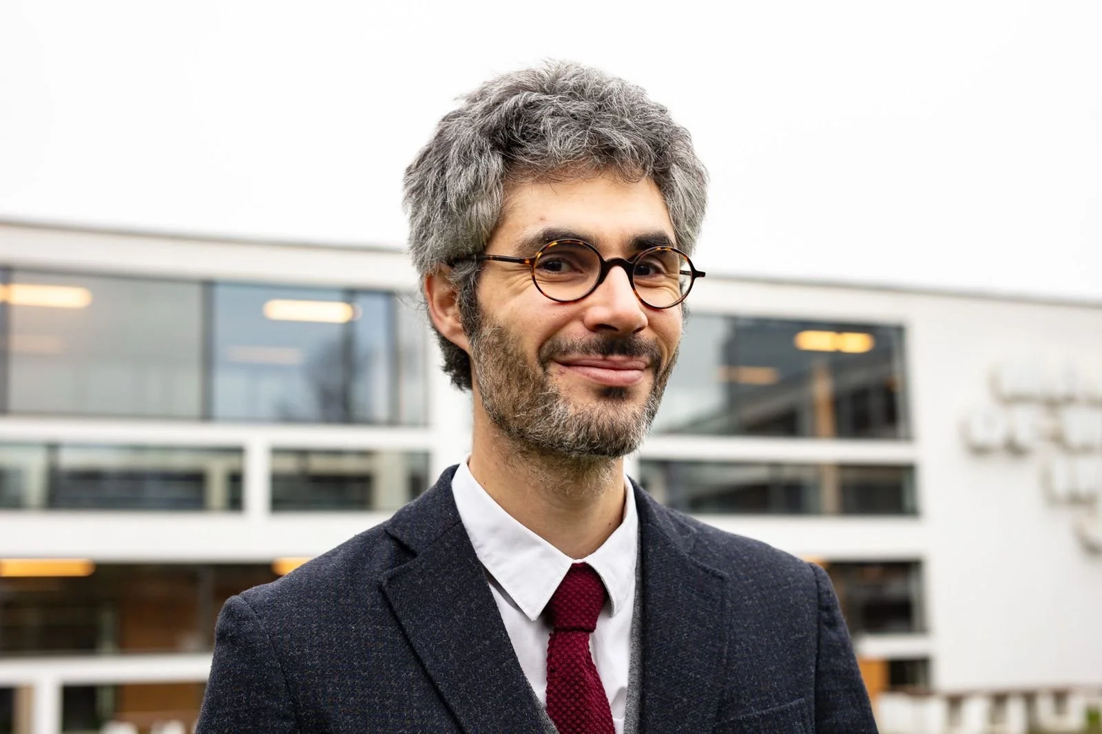 A middle-aged man with gray hair, beard, and glasses smiling outdoors in front of a modern office building.