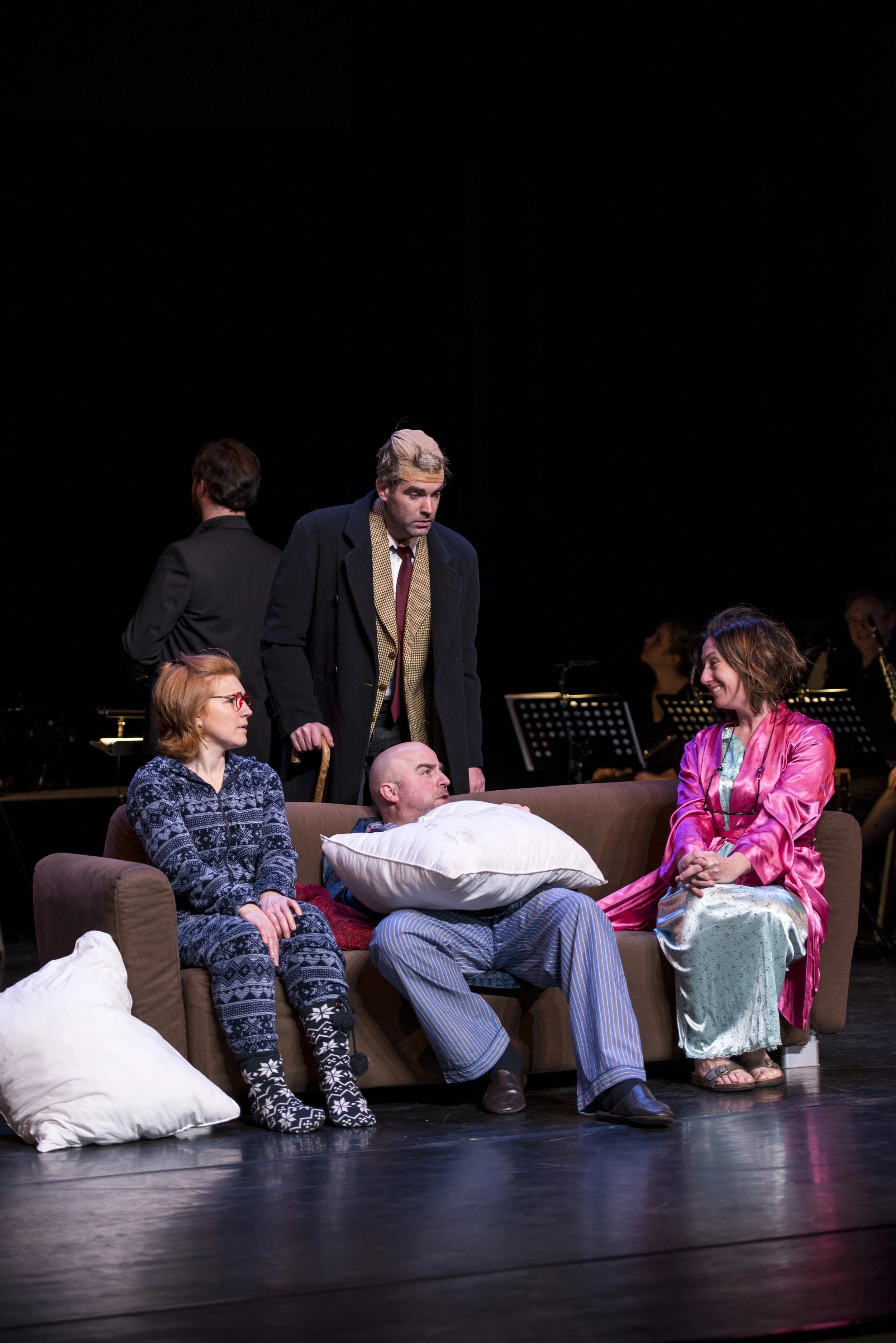 Actors performing a scene on stage, with some sitting on a sofa and others standing around, depicting a theatrical play.