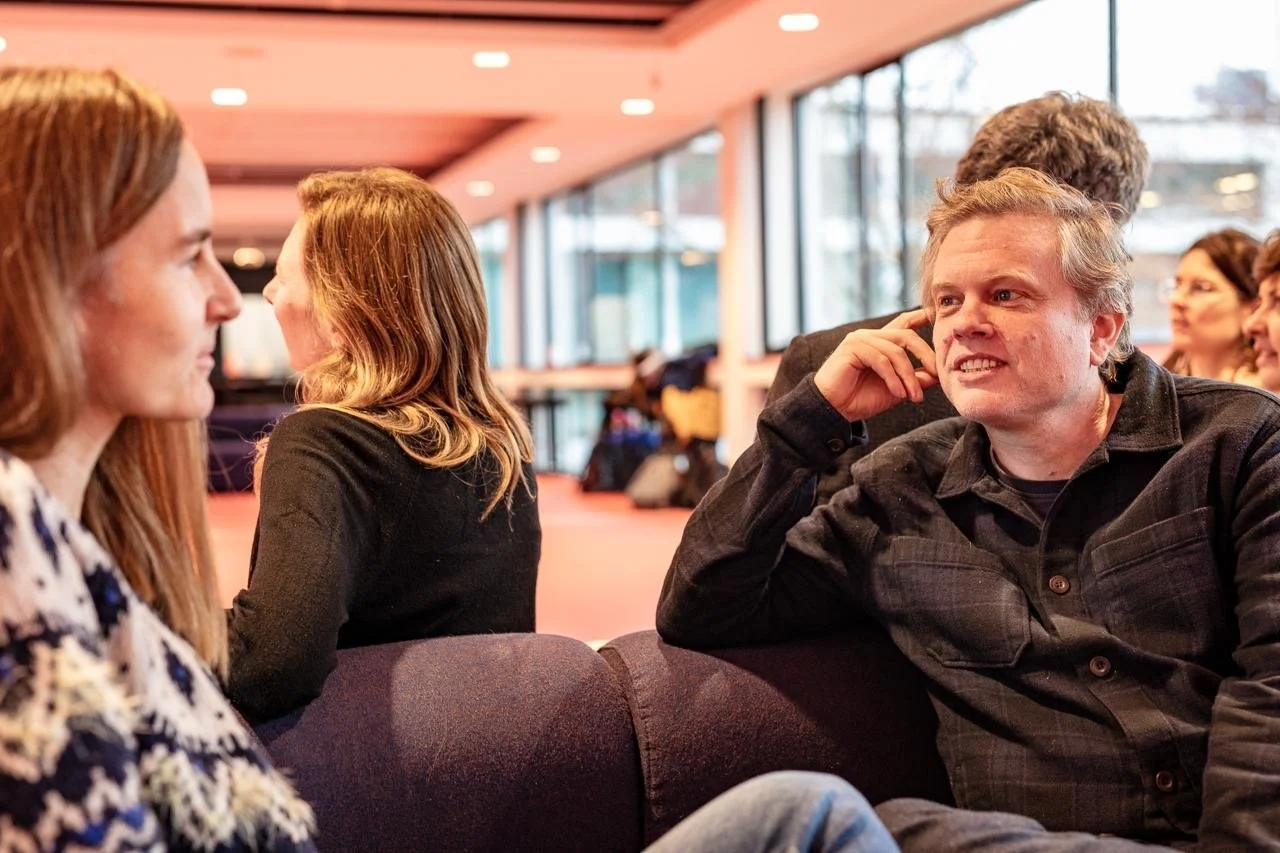 People sitting in a lobby engaged in conversation, with large windows and modern decor in the background.