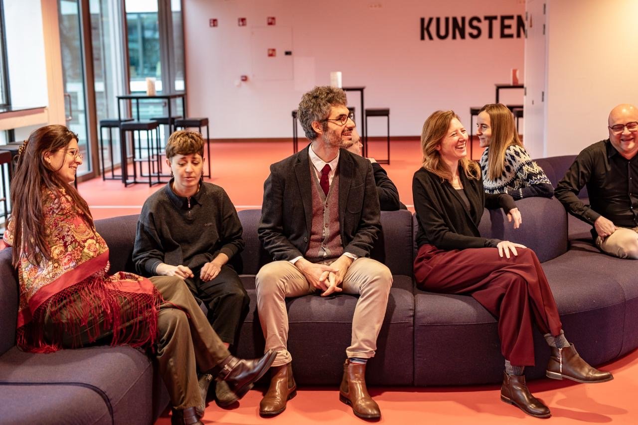 Group of six people sitting on a purple couch, smiling and laughing in a modern, brightly lit room with large windows and a pink floor, with the word 'KUNSTEN' on the wall.