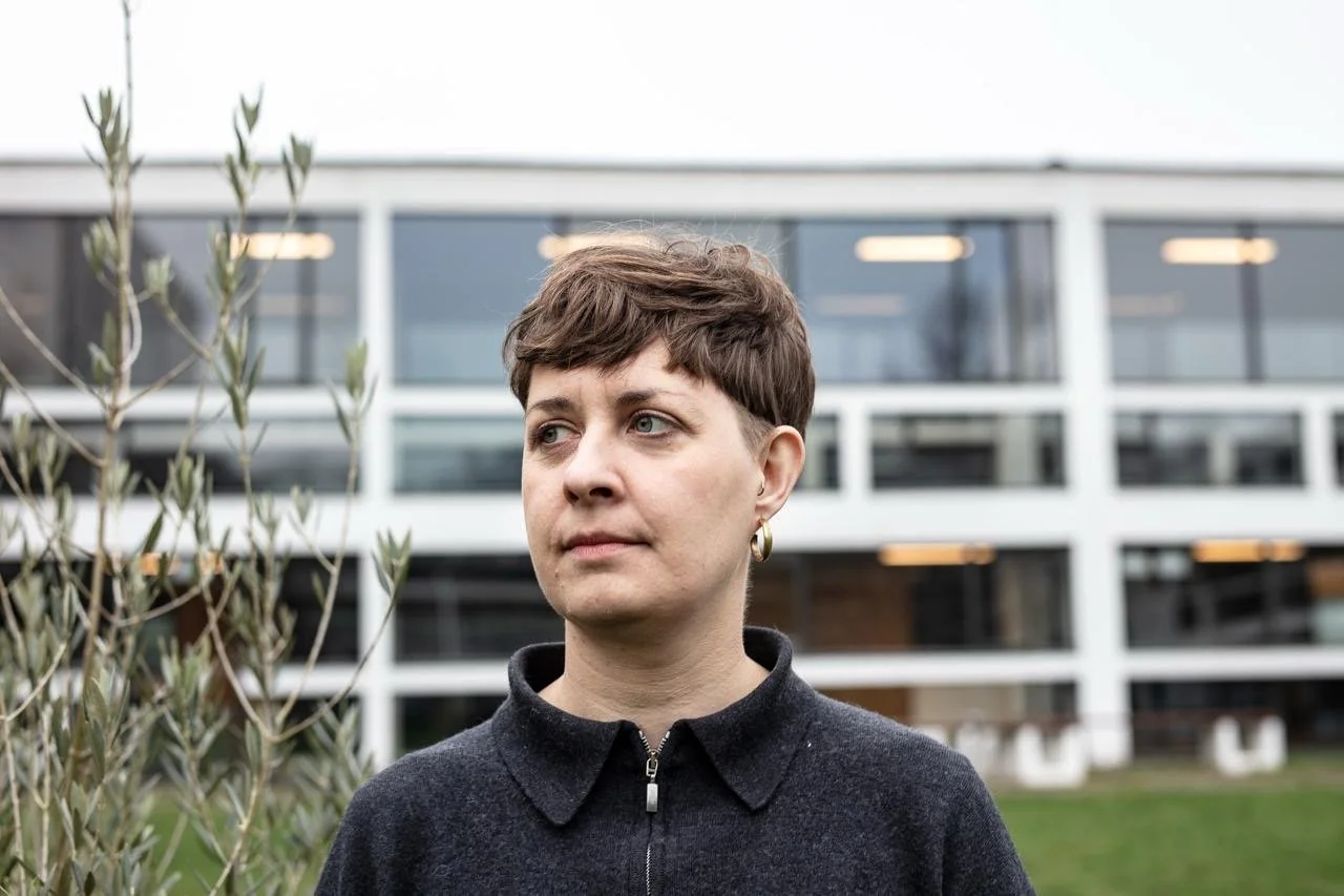 A woman with short brown hair and hoop earrings stands outdoors in front of a modern building with large windows, wearing a dark zip-up jacket.