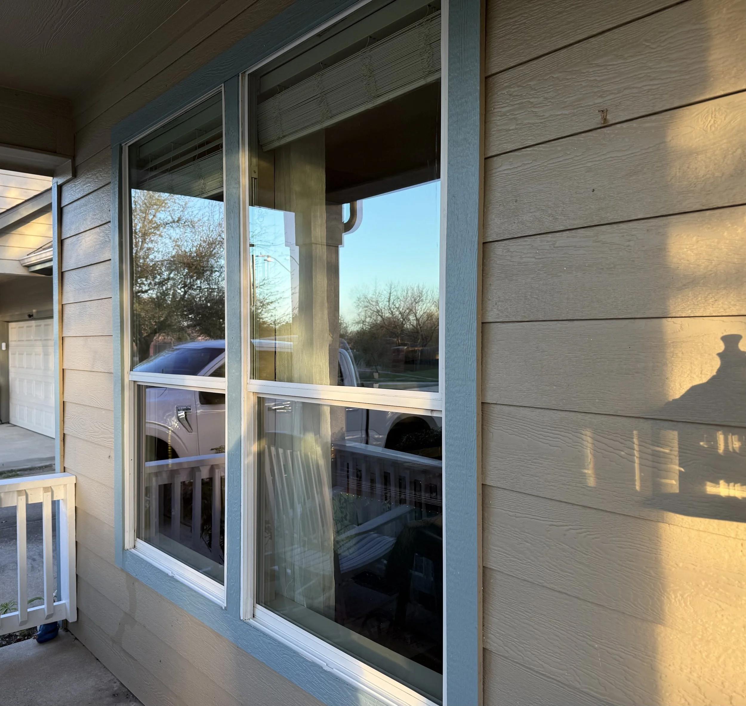 Exterior of a house with beige horizontal siding, a large window reflecting a truck, porch, and sunset lighting.