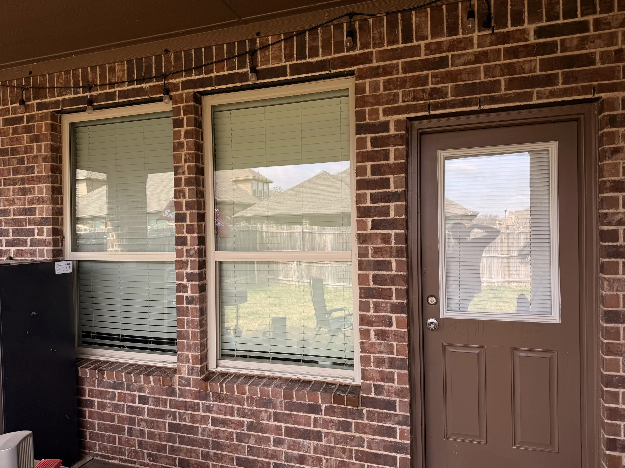 Exterior view of a brick house showing two large windows with closed white blinds and a brown door with a glass window, reflecting the backyard and neighboring rooftops.