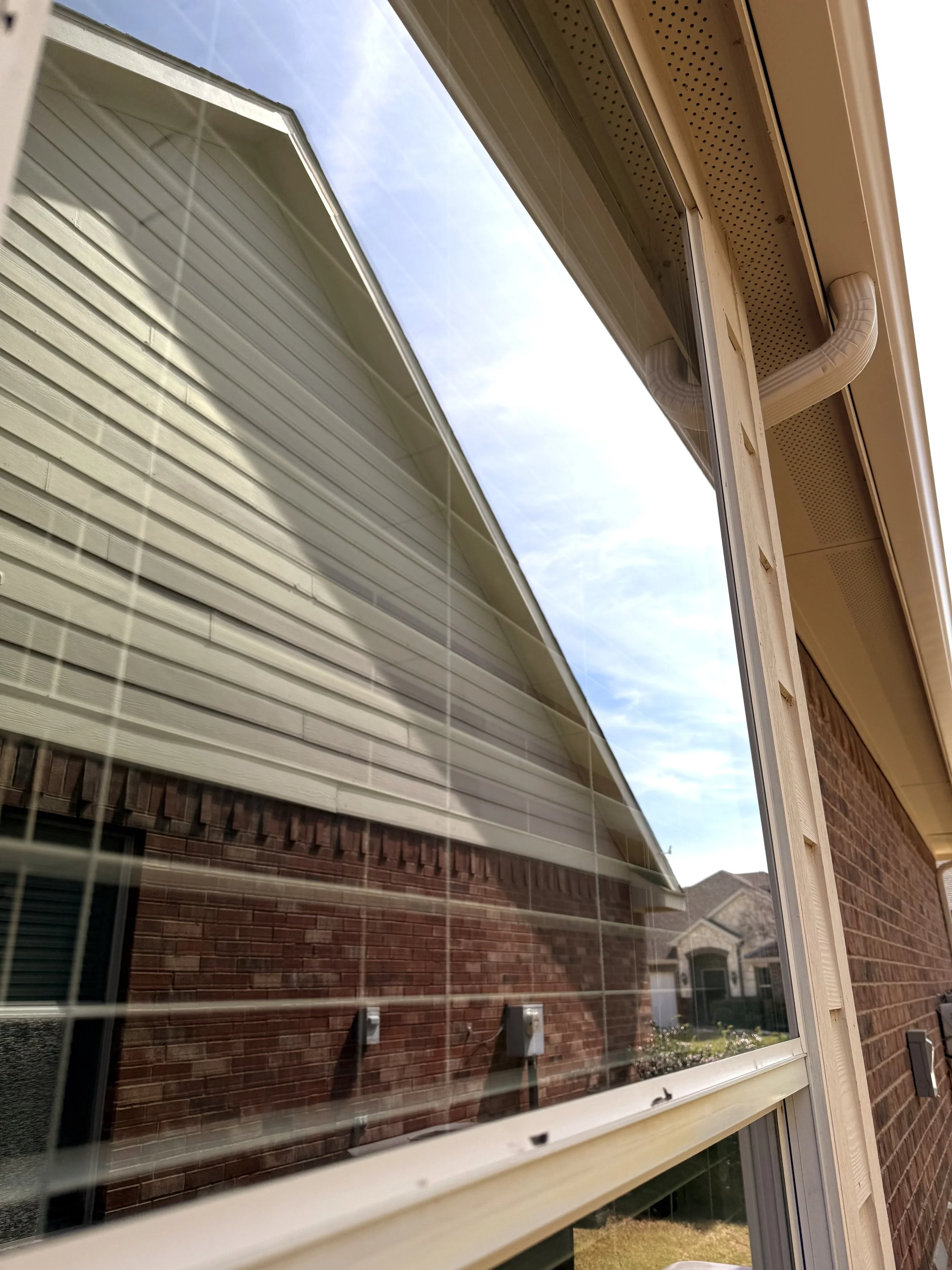 Close-up of a house window with blinds, reflecting the brick wall of a neighboring house and the sky.