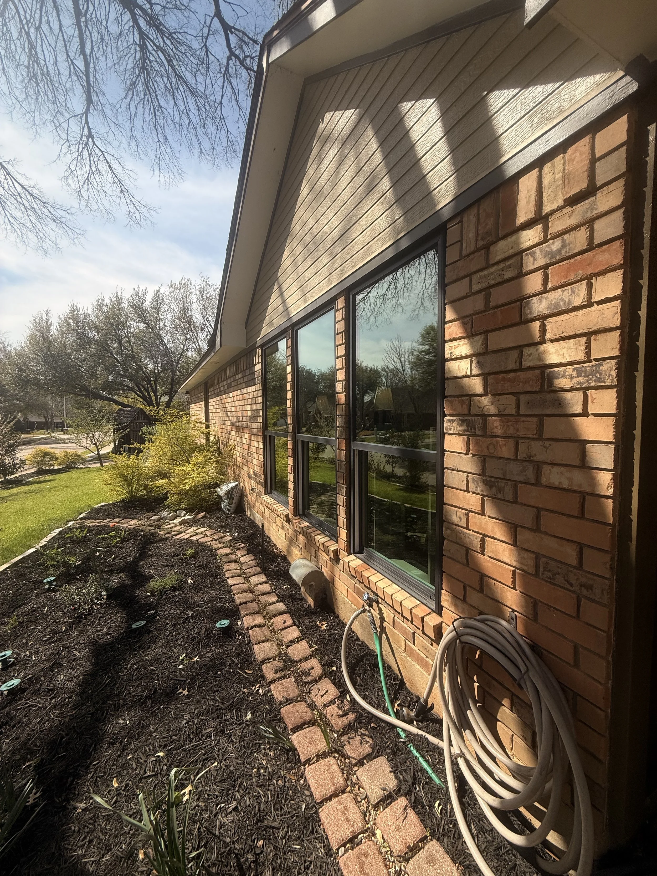 Side view of a brick house with multiple windows, a garden bed with soil and plants, and a coiled garden hose on the brick pathway, under a tree with leafless branches, during daylight.