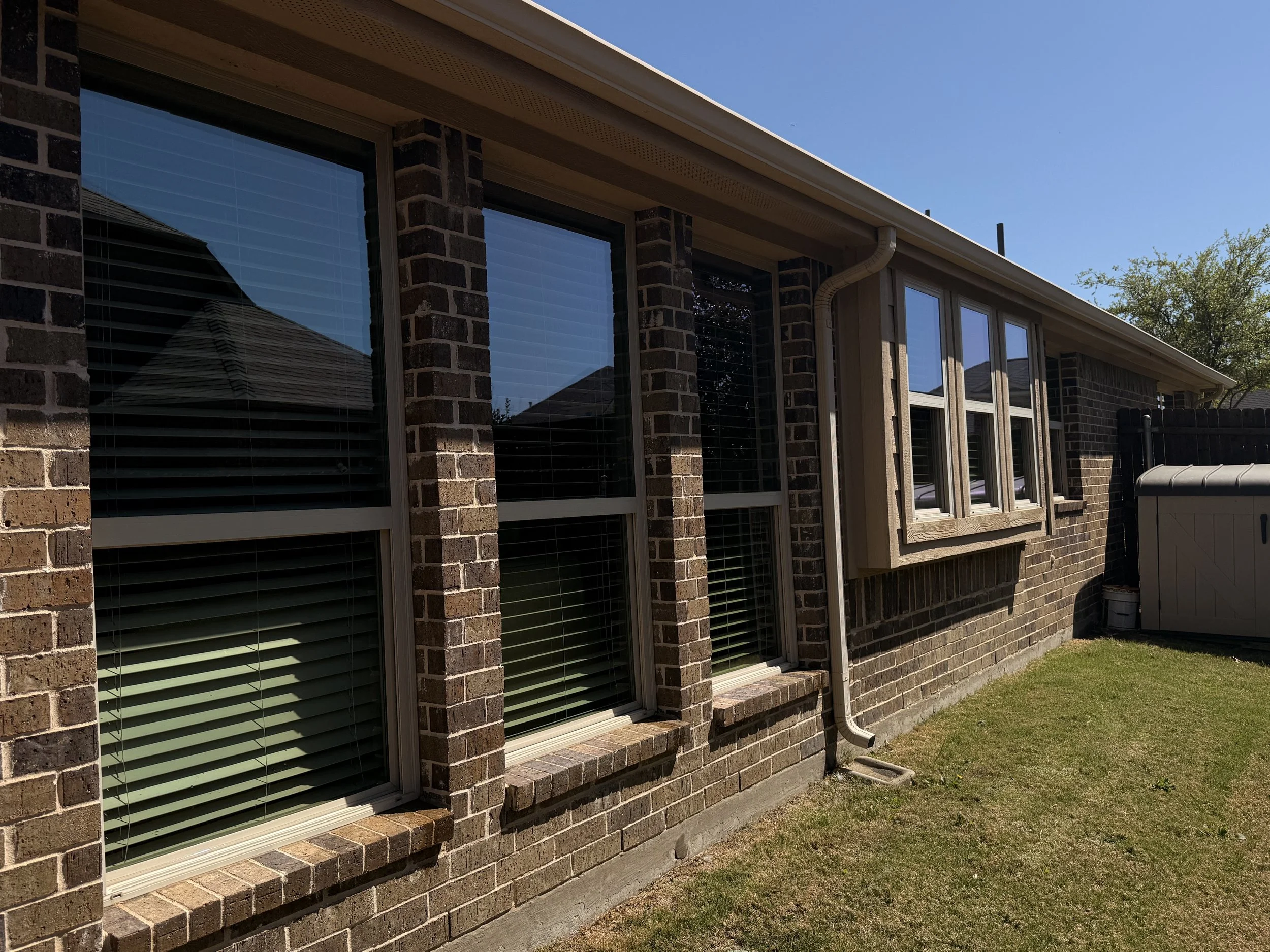 The exterior of a brick house with multiple windows, some with blinds, and a small grassy yard. A gutter runs along the roof edge, and a storage shed is visible in the back corner.