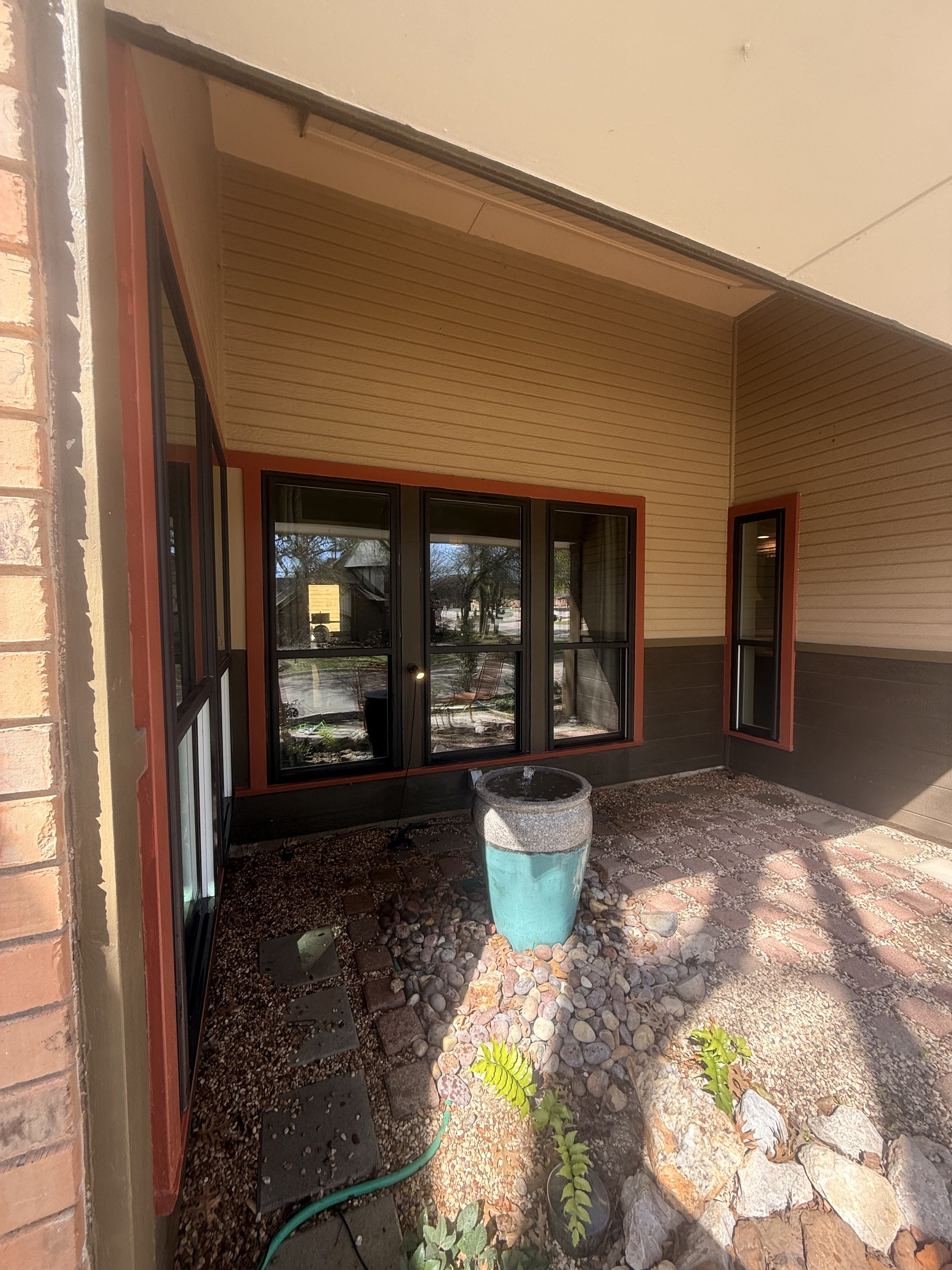 A small outdoor porch area with a water fountain and a few plants, enclosed by a brick wall, with windows and beige siding on the house.
