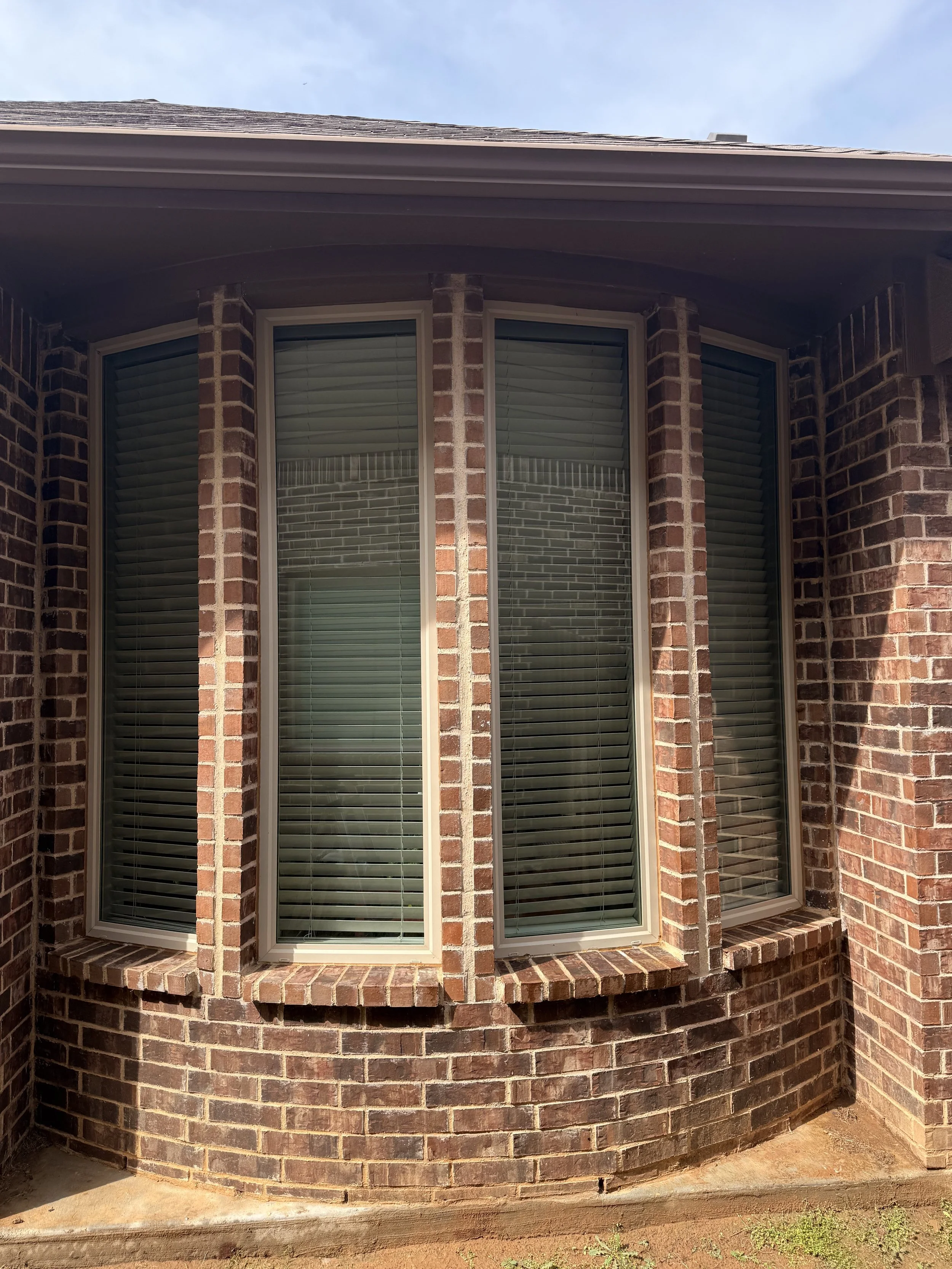 Brick house with three large windows featuring beige trim, window blinds, and a curved brick wall.