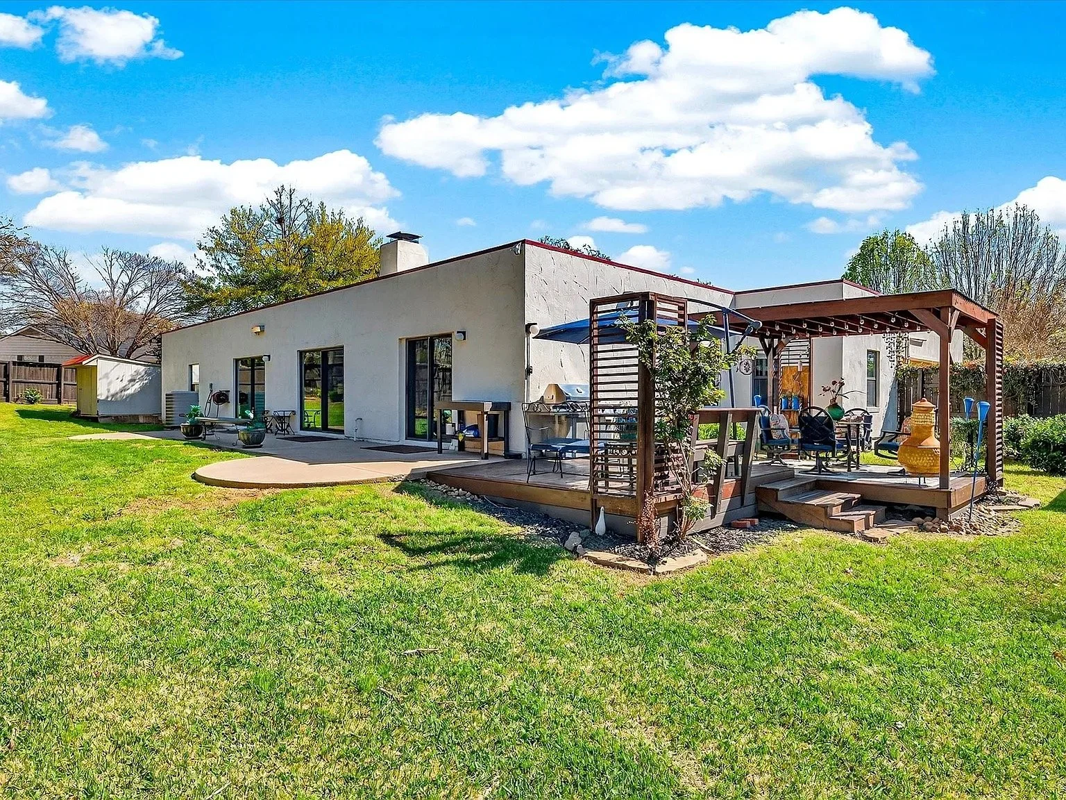 Backyard with a grassy lawn, concrete patio, and a wooden deck with outdoor seating under a pergola, against a white stucco house with sliding glass doors, under a blue sky with scattered clouds.