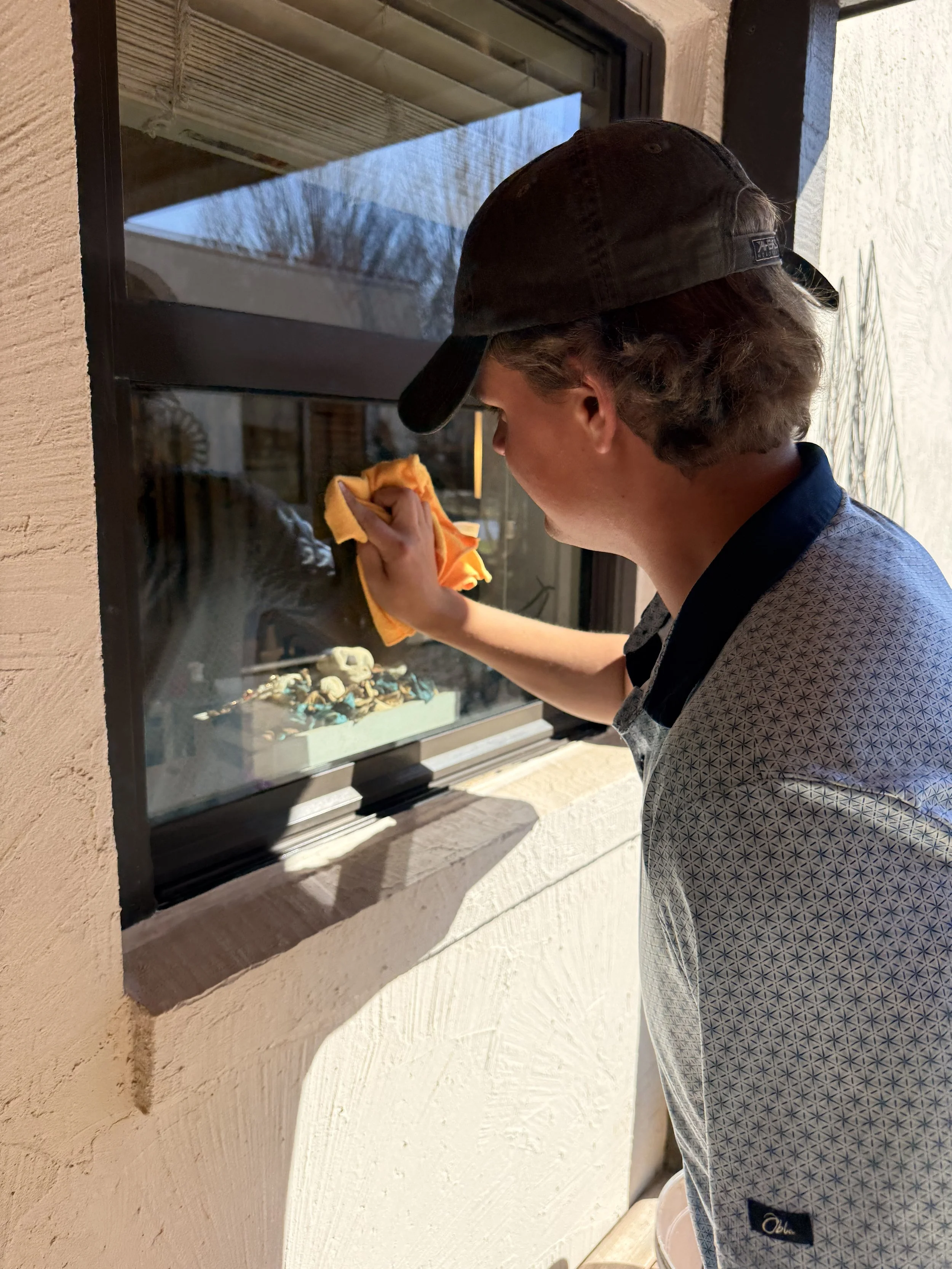 A person wearing a black cap and patterned shirt is cleaning a window with an orange cloth, viewing the inside of a room with various objects, including skull figurines, on a table.