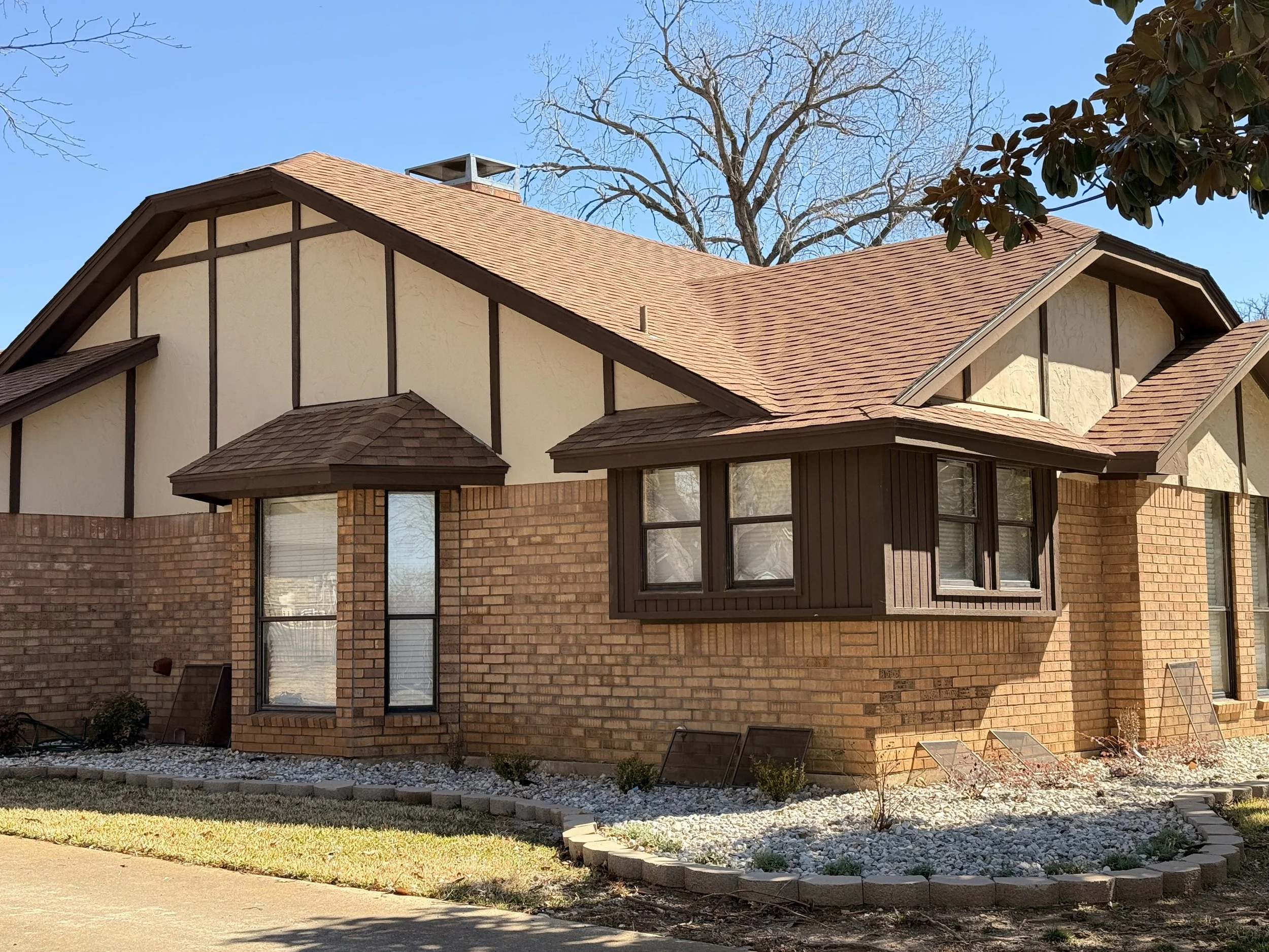 A two-story house with brick and white siding exterior, brown roof, and several windows, including a bay window. The house is surrounded by a small landscaped yard with rocks and a brick border, and some trees are visible in the background.