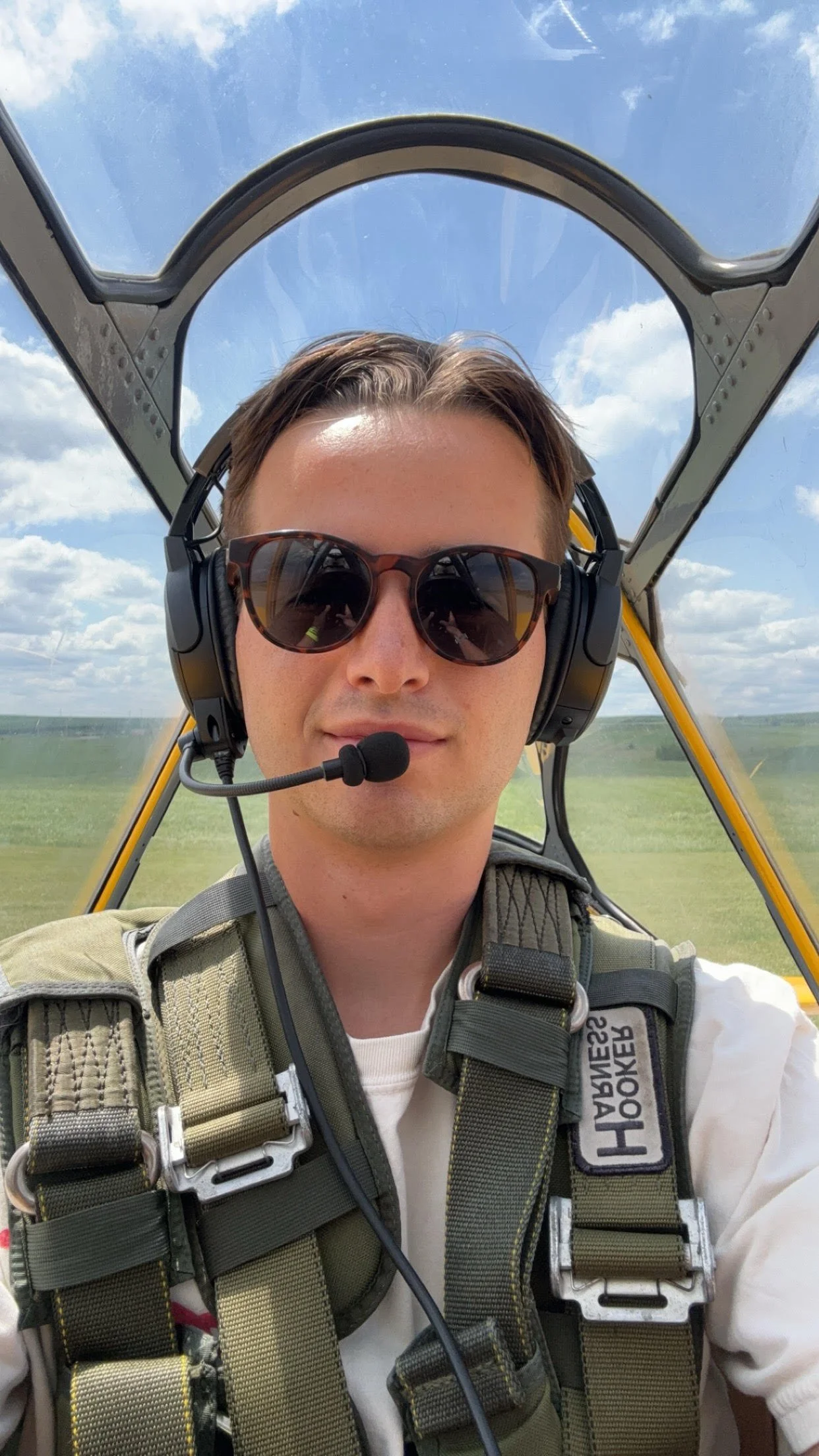 A man wearing sunglasses and a headset taking a selfie inside a small aircraft cockpit, with blue sky and clouds visible through the canopy.