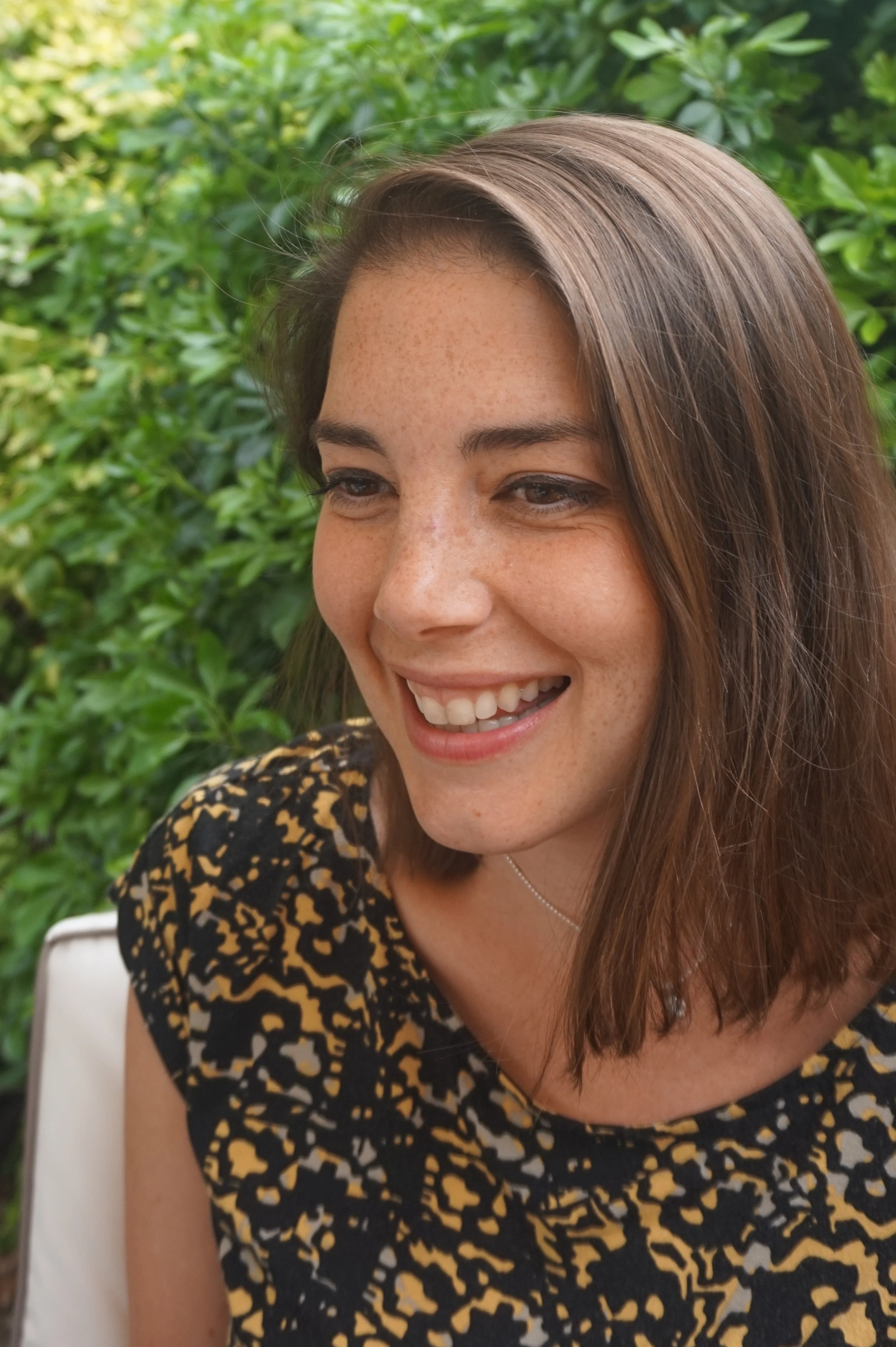 Close-up of a smiling woman with medium-length brown hair, wearing a black and yellow patterned top, outdoors with green foliage in the background.