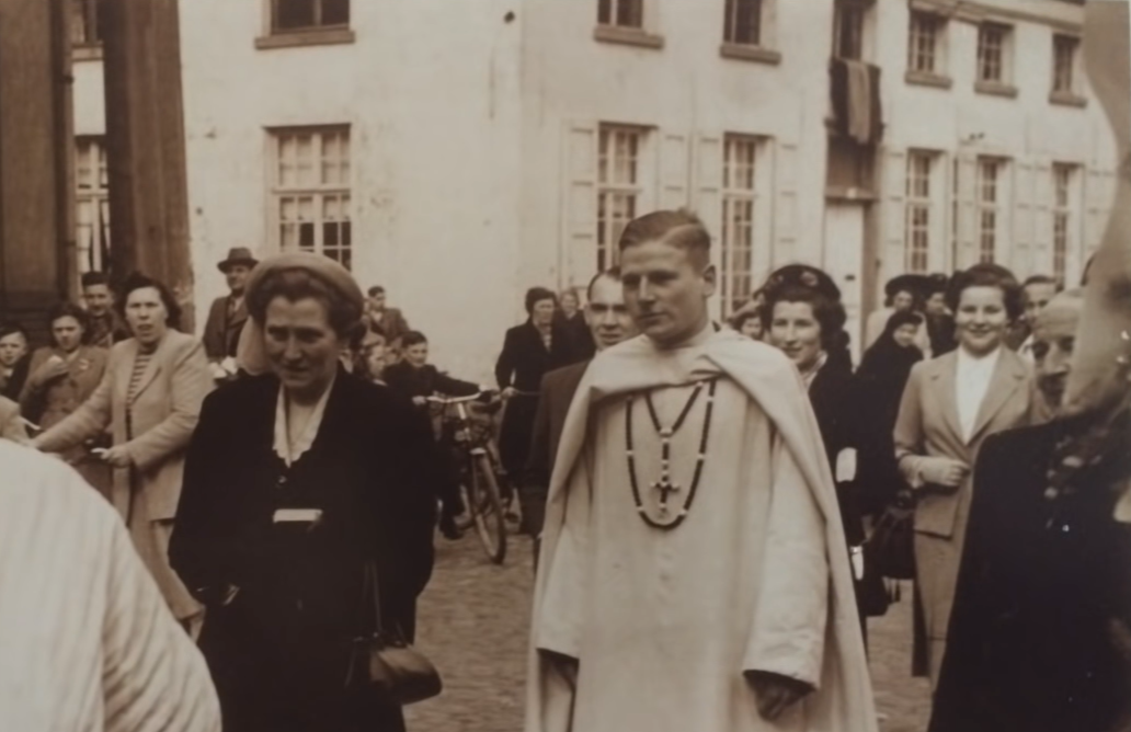 Photo en noir et blanc montrant un groupe de personnes debout à l'extérieur devant un immeuble, avec un jeune homme en robe cérémoniale ou cape avec des colliers de perles et une croix, entouré de femmes et d'hommes d'apparence vintage.