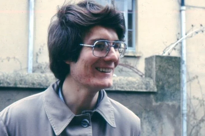 Femme portant des lunettes et un trench beige, souriant, à l'extérieur avec un mur en pierre en arrière-plan.