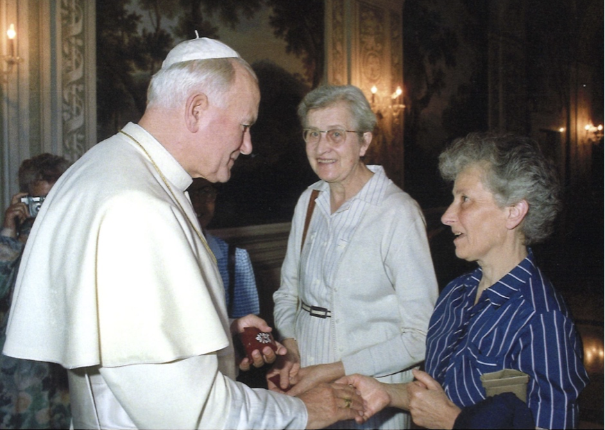 Le pape François échange une poignée de main avec deux femmes lors d'une rencontre officielle, dans un lieu décoré avec des peintures murales et un éclairage chaleureux.