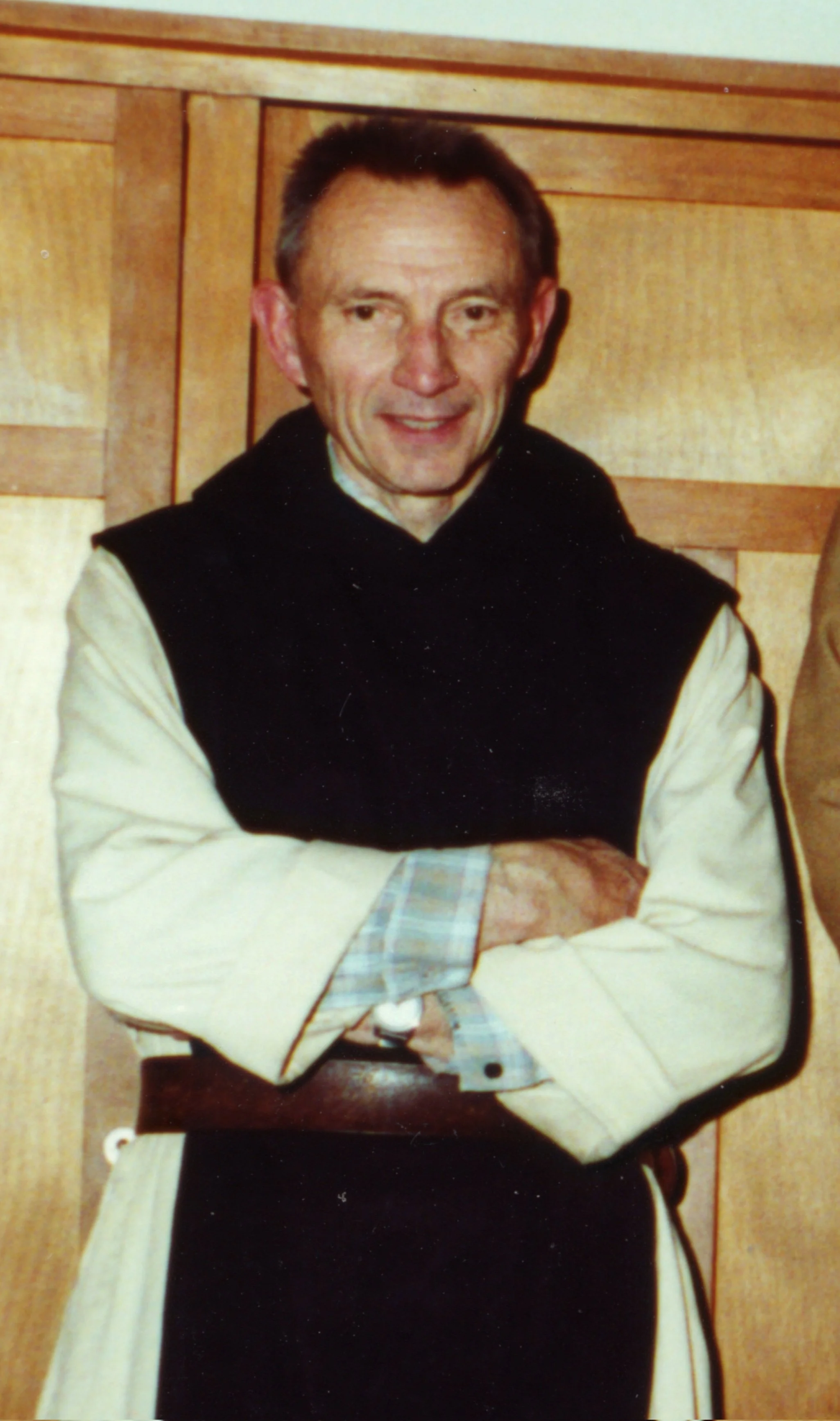 Un homme d'âge moyen avec des cheveux courts, souriant, portant un pull noir et une chemise beige avec un motif à carreaux. Il est assis à une table en bois devant un fond en bois clair.