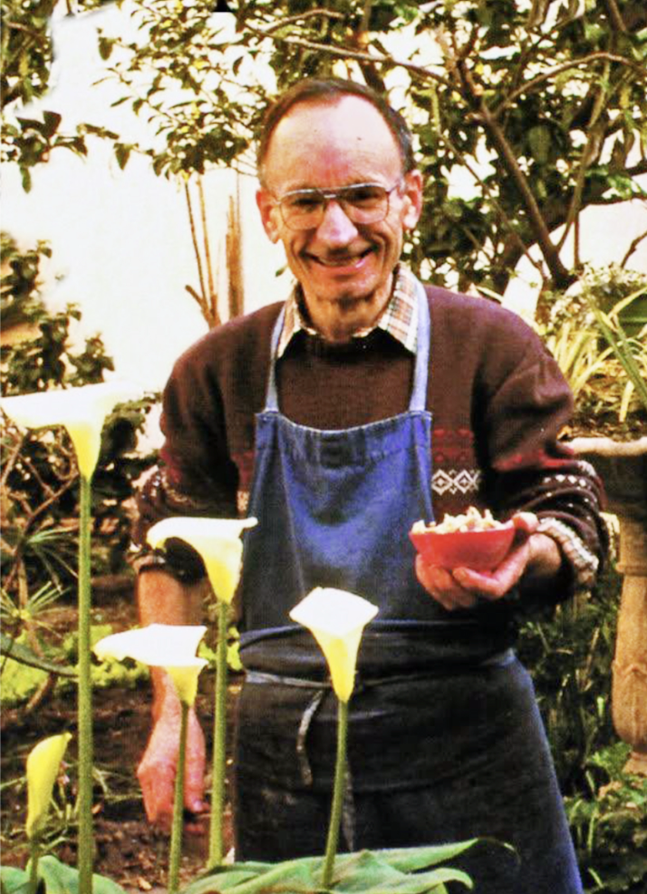 Un homme âgé souriant, portant une chemise à carreaux, un pull marron, un tablier bleu, se tenant dans un jardin entouré de plantes, tenant un bol de nourriture.