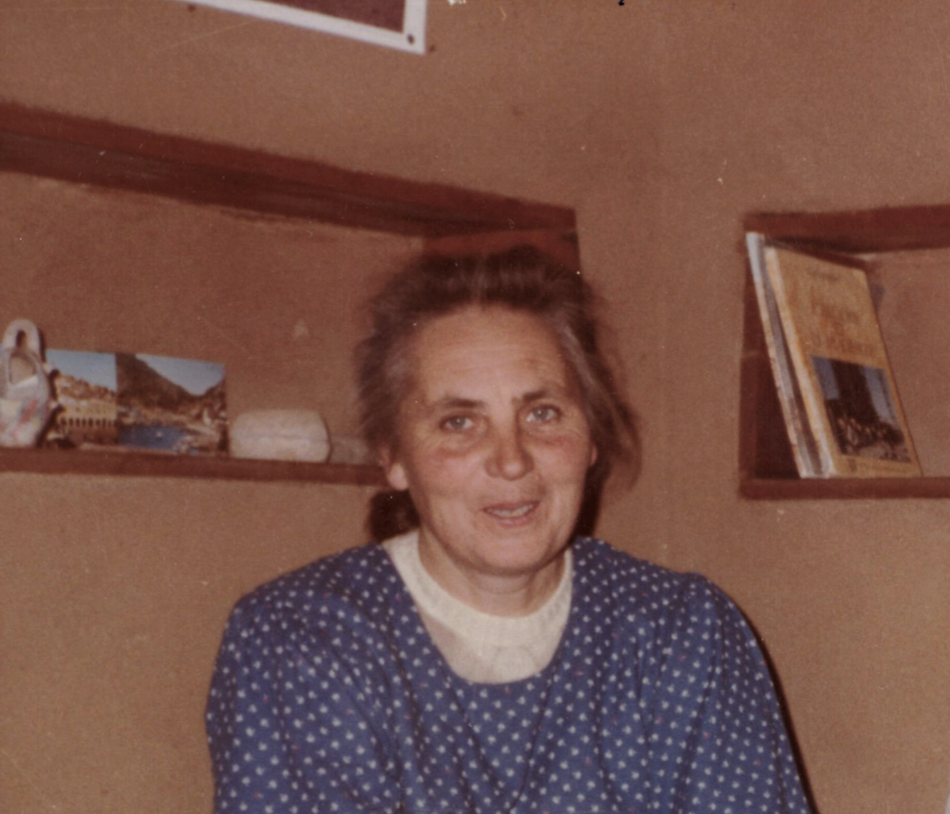 Une femme âgée souriante, portant un pull à pois bleu et blanc, dans une pièce avec des étagères en bois contenant des livres et quelques objets.