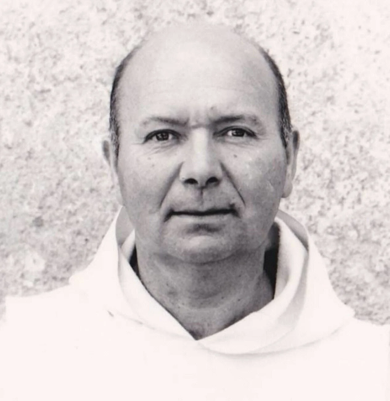 Photo en noir et blanc d'un homme avec une tête chauve, portant un hoodie blanc, avec un fond de texture rocheuse ou granuleuse.