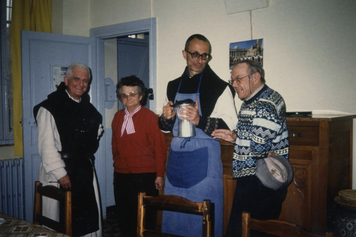 Cinq personnes debout dans une pièce, trois hommes et deux femmes, posant pour une photo. L'un des hommes porte un tablier bleu et tient une cafetière, les autres sourient.