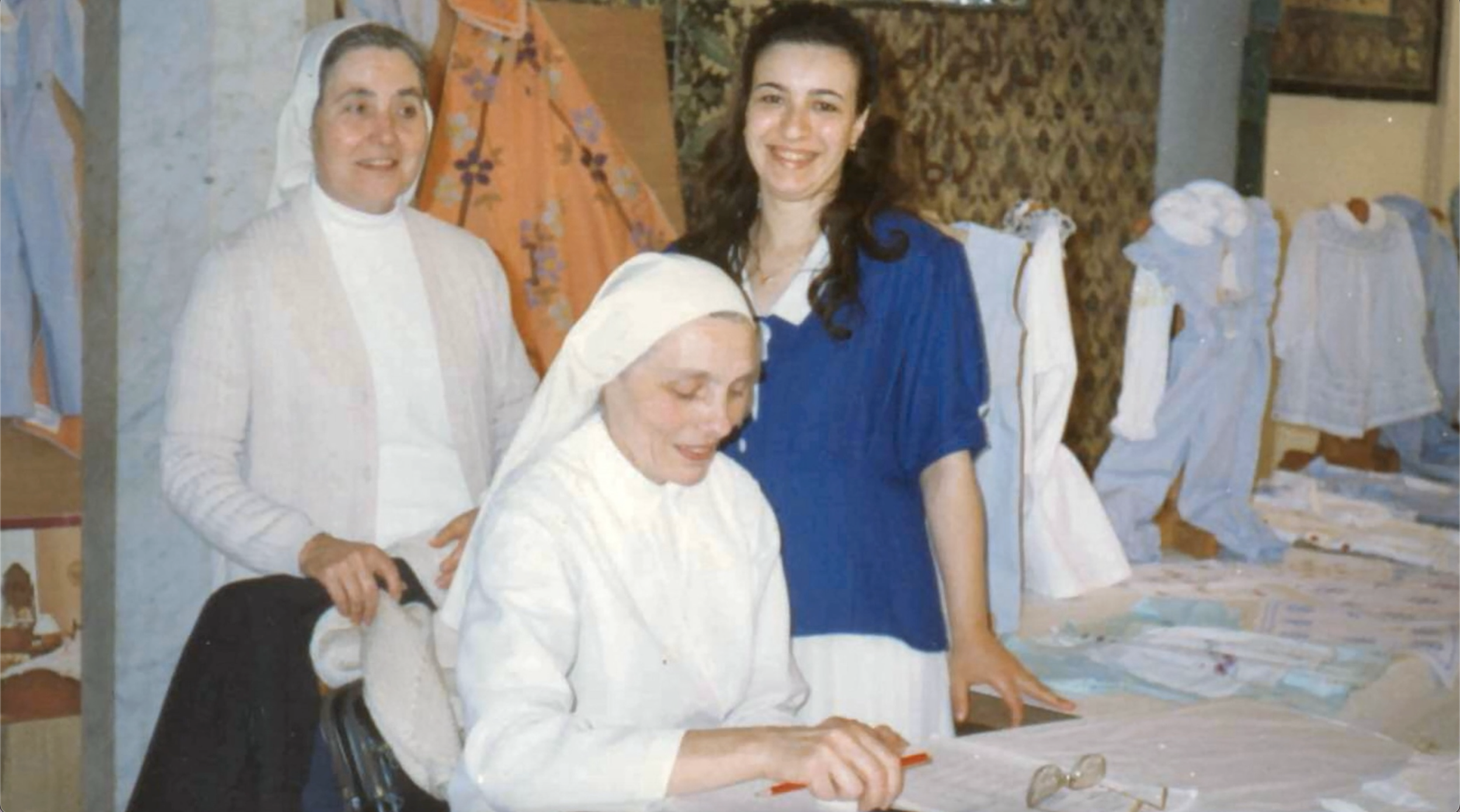 Quatre femmes, dont deux en uniformes de travail, autour d'une table avec des vêtements ou des tissus. La scène semble se dérouler dans un atelier ou un espace de couture.