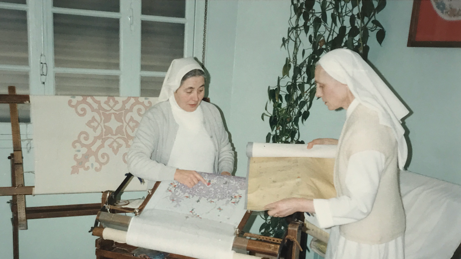 Deux femmes habillées en habits de religieuses, l'une travaillant sur un métier à tisser avec un tableau de broderie, l'autre montrant un rouleau de papier.