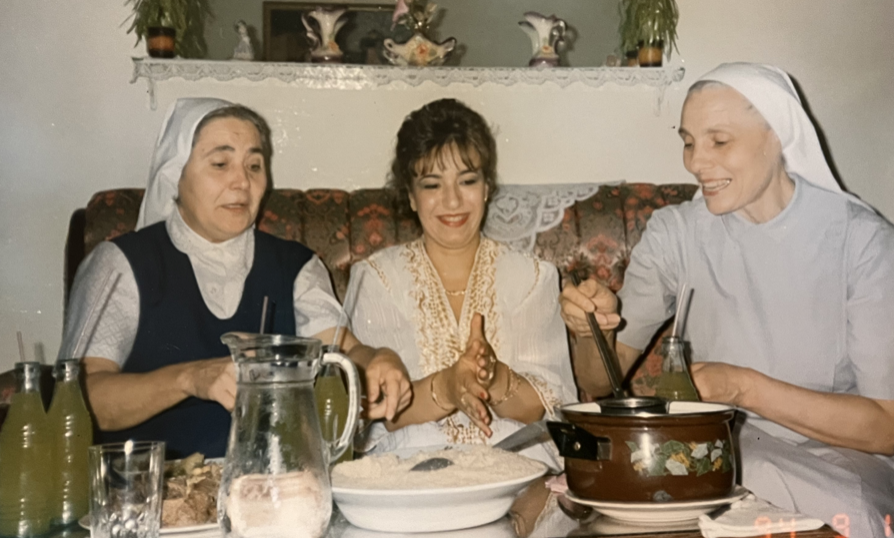 Trois femmes, dont deux religieuses en uniforme, autour d'une table partageant un repas, souriantes.