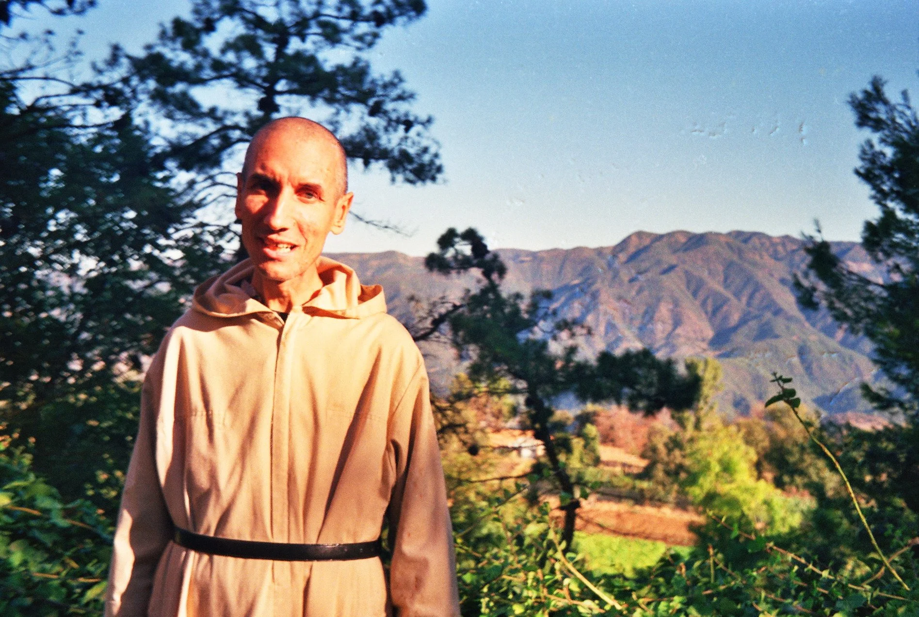 Une personne en robe beige devant un paysage de montagnes et de forêt, avec un ciel clair.