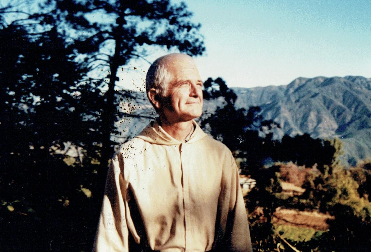 Un homme âgé, vêtu d'une robe beige, se tient dans un paysage montagneux avec des arbres et une montagne en arrière-plan, regardant vers la lumière du soleil.