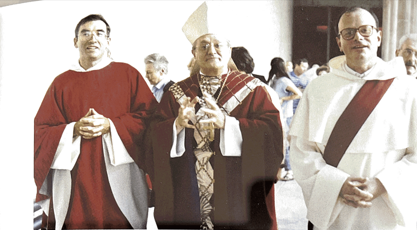 Trois personnes en tenue académique lors d'une cérémonie. La personne au centre porte une robe de graduation et un chapeau cardinalice, la personne à gauche porte une toge rouge, et celle à droite porte une toge blanche avec une croix diagonale rouge.