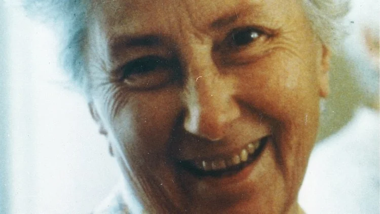 Photo d'une femme âgée souriante avec des rides et des cheveux gris ou blancs, éclairée doucement.