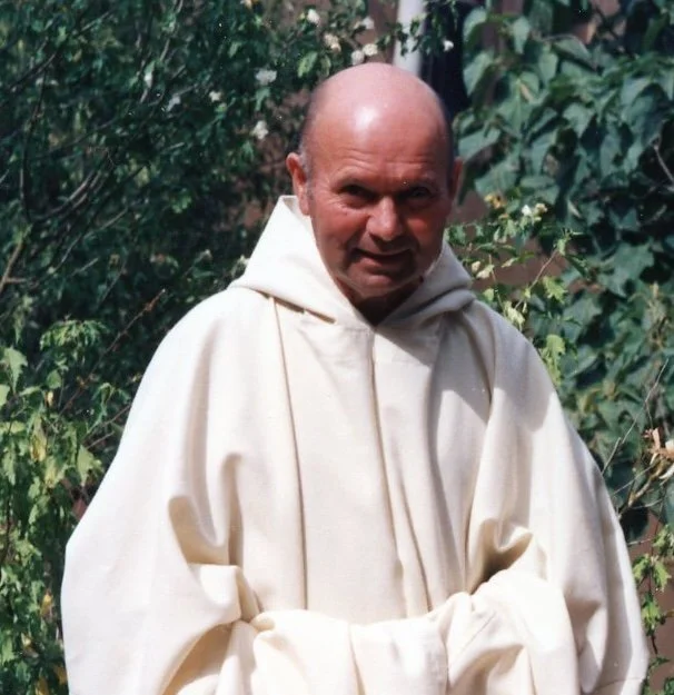 Un homme vêtu d'une robe blanche, debout devant des plantes et des feuillages