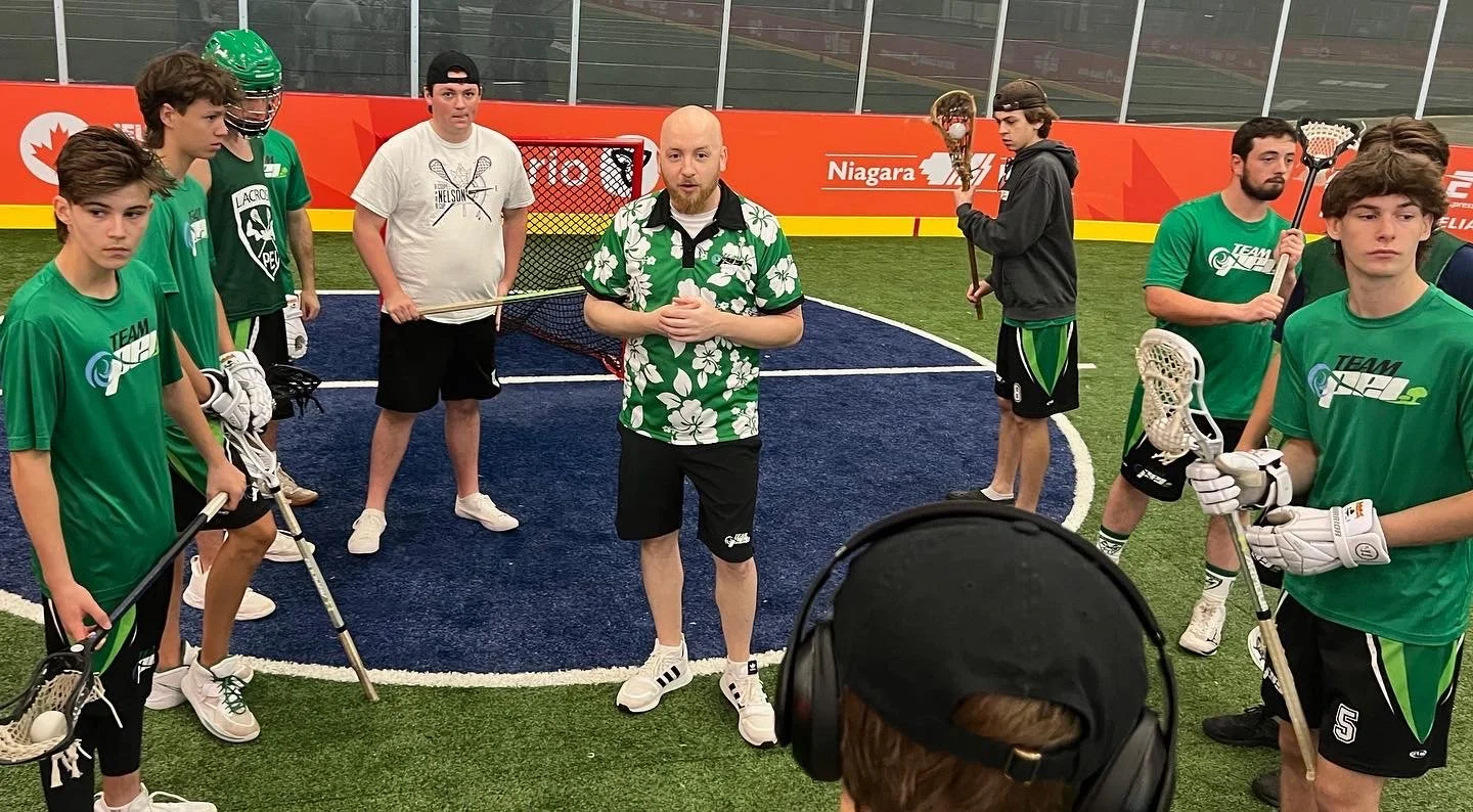 Lacrosse team practice on an indoor field with coach giving instructions, players holding lacrosse sticks, wearing green jerseys and gloves.