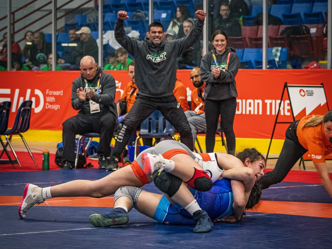 Two female wrestlers in a match on the mat, with one wrestler pinning the other down. Audience and officials watch from the background, with some people standing and cheering.