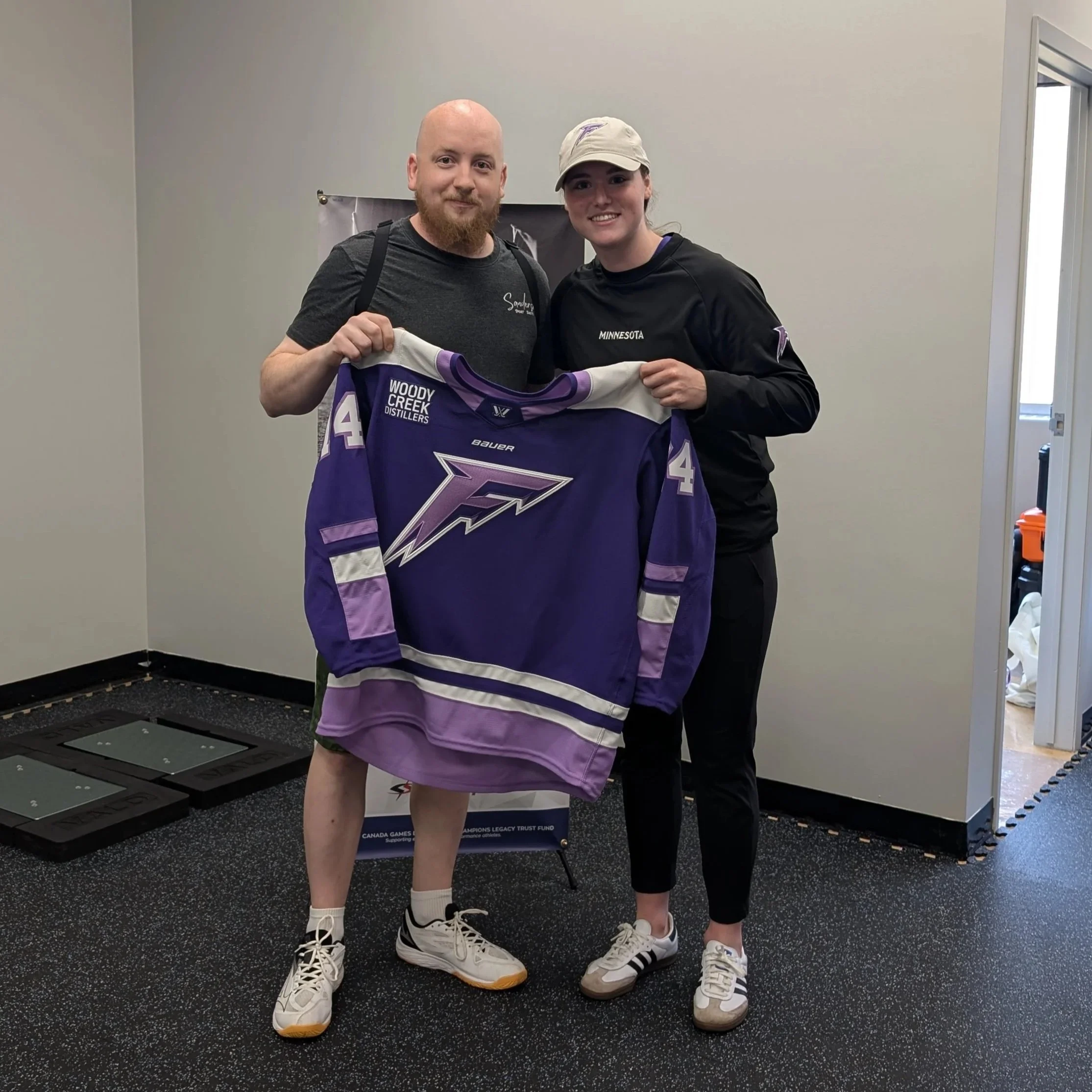 A man and woman stand together holding a purple hockey jersey with the number 44 and a logo, inside a room with a display of scales and a poster in the background.