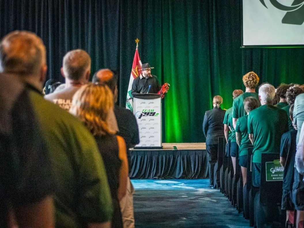 A group of people standing and facing a stage where a man is giving a speech at a podium. The background is decorated with a black curtain, a flag, and green lighting.