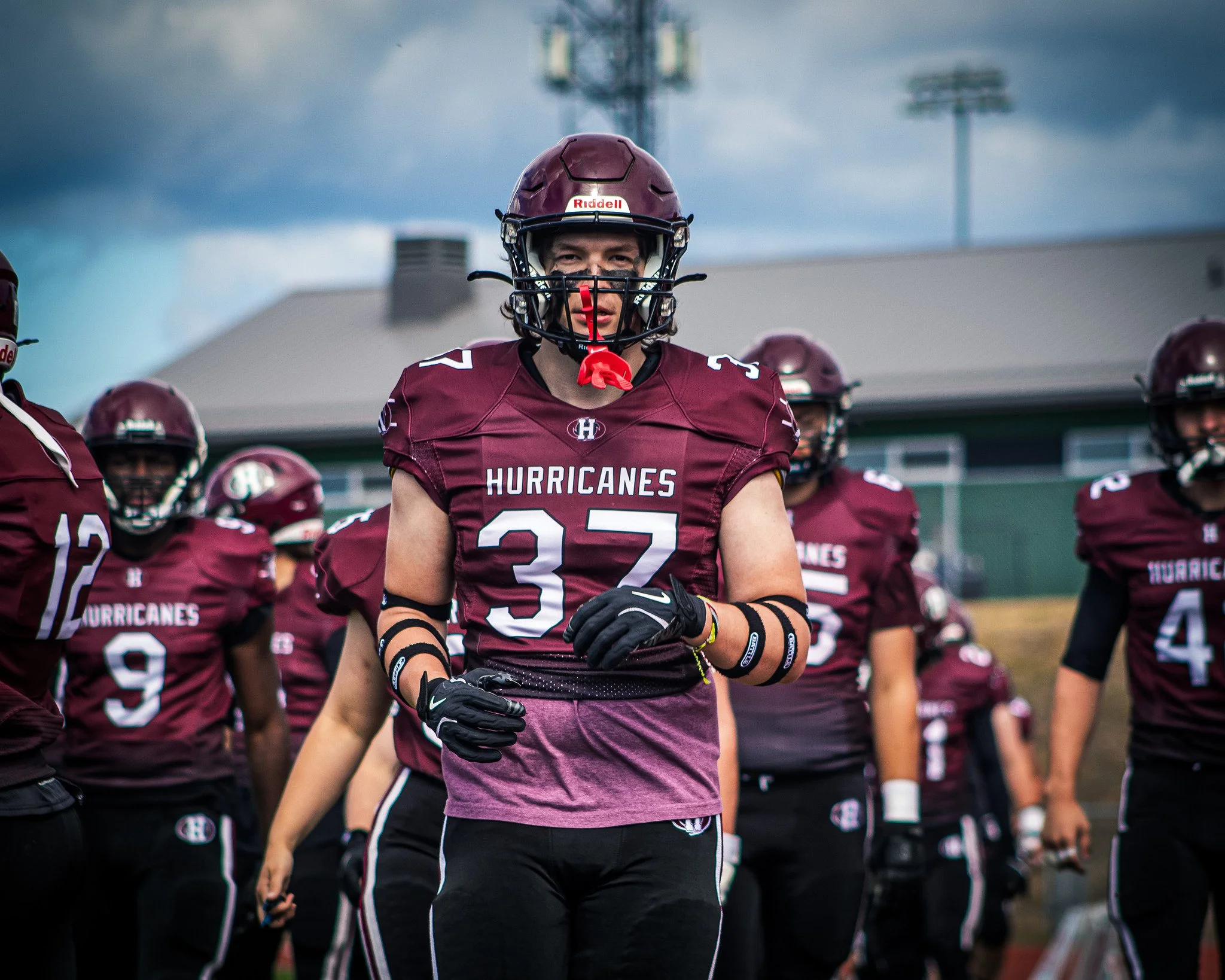 A group of football players wearing maroon jerseys and helmets, standing on a field with a sports facility and cloudy sky in the background.