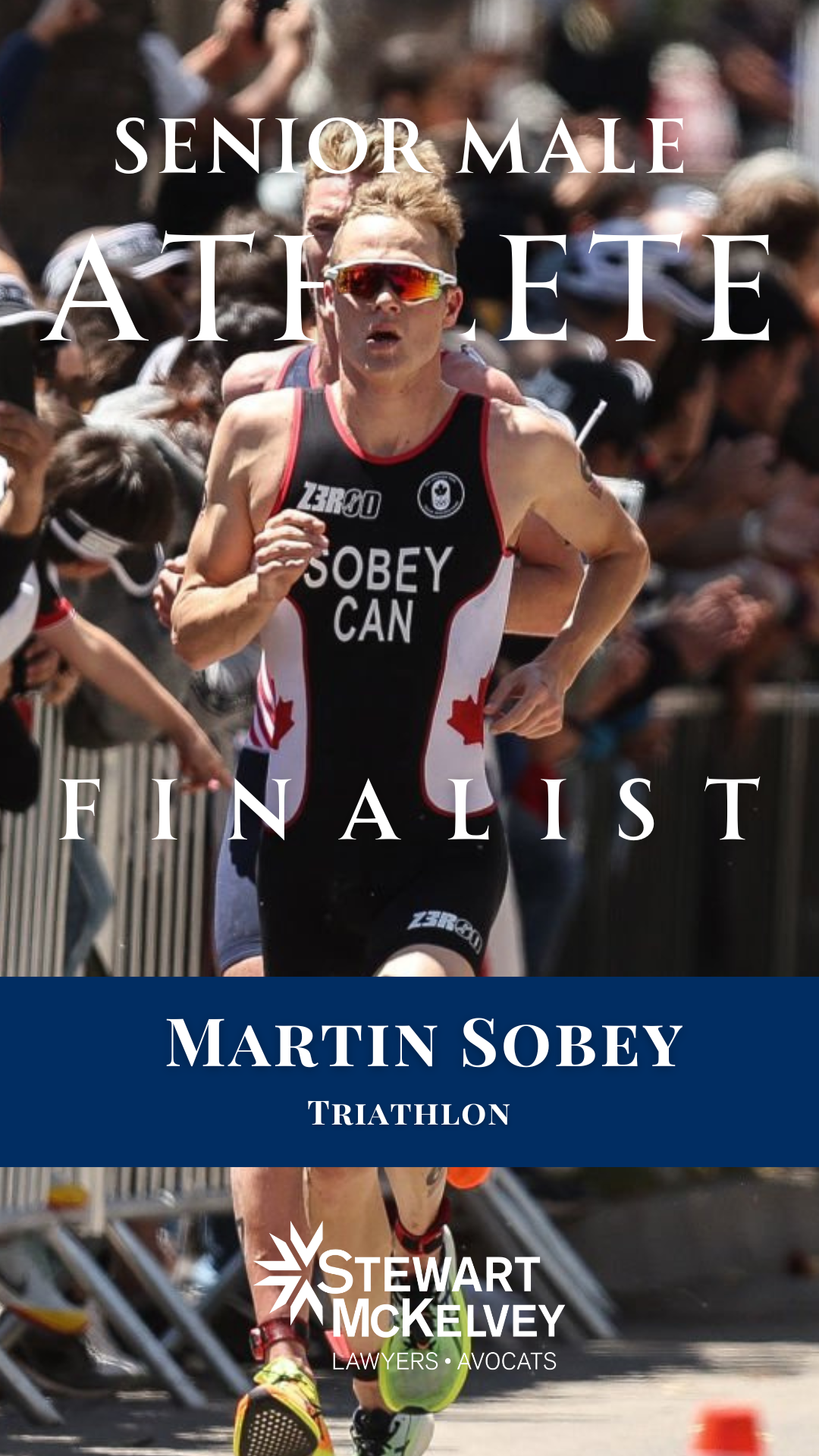 A male triathlete running in a race with a crowd of spectators in the background. He is wearing sunglasses, a black triathlon suit with red and white accents, and the text 'SOBEY CAN' on the front.