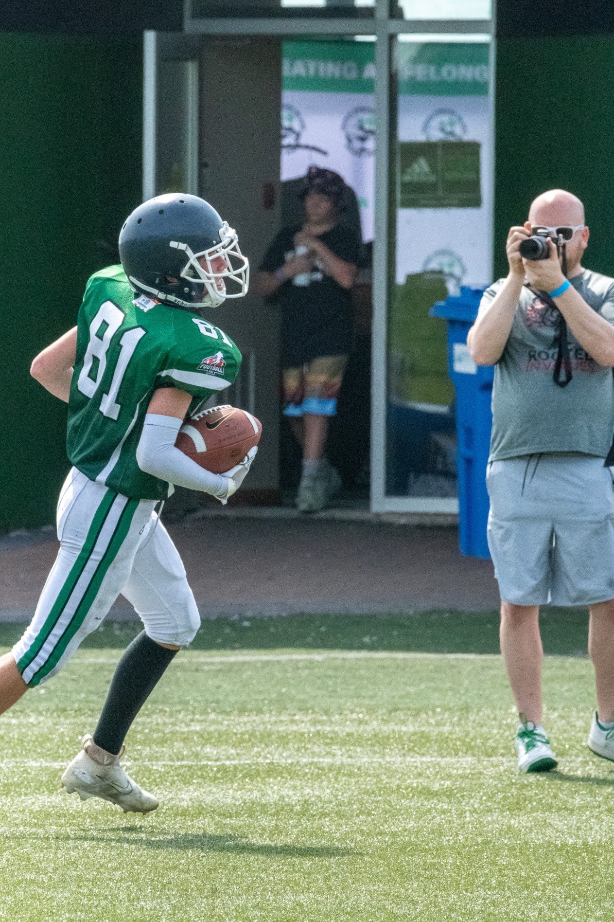 A young football player in a green uniform holding a football runs on the field, with a photographer capturing the moment nearby, and people in the background near the entrance.