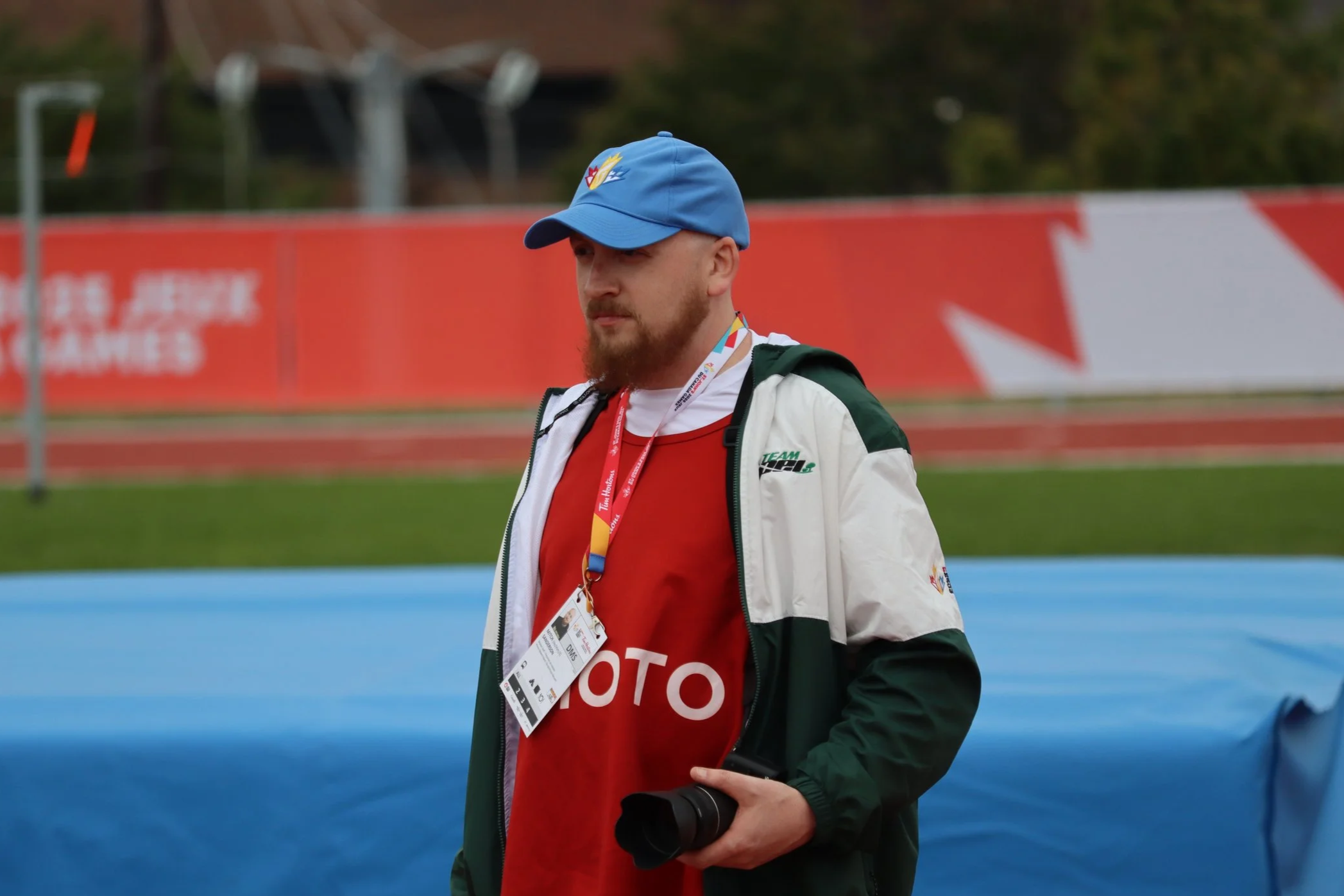 A man wearing a blue cap, a red shirt with the word 'MOTO' on it, and a white jacket stands outdoors on a sports field, holding a camera in his right hand, with a colorful lanyard and badge around his neck.
