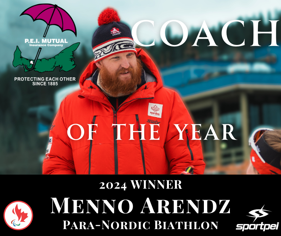 A man with a red beard and wearing a red winter jacket, a black and white headband, and a black knit hat with a red pompom, standing outdoors. The background shows a building and cloudy sky.