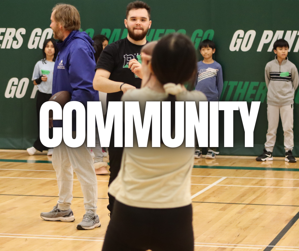 People gathered in a gymnasium for a community event, with a large green backdrop that reads 'GO PANTHERS' and 'GO PANTHERS GO.' A man with a beard and wearing a black t-shirt is in the center, interacting with a young girl with glasses. Several children are standing in the background, some holding green papers.
